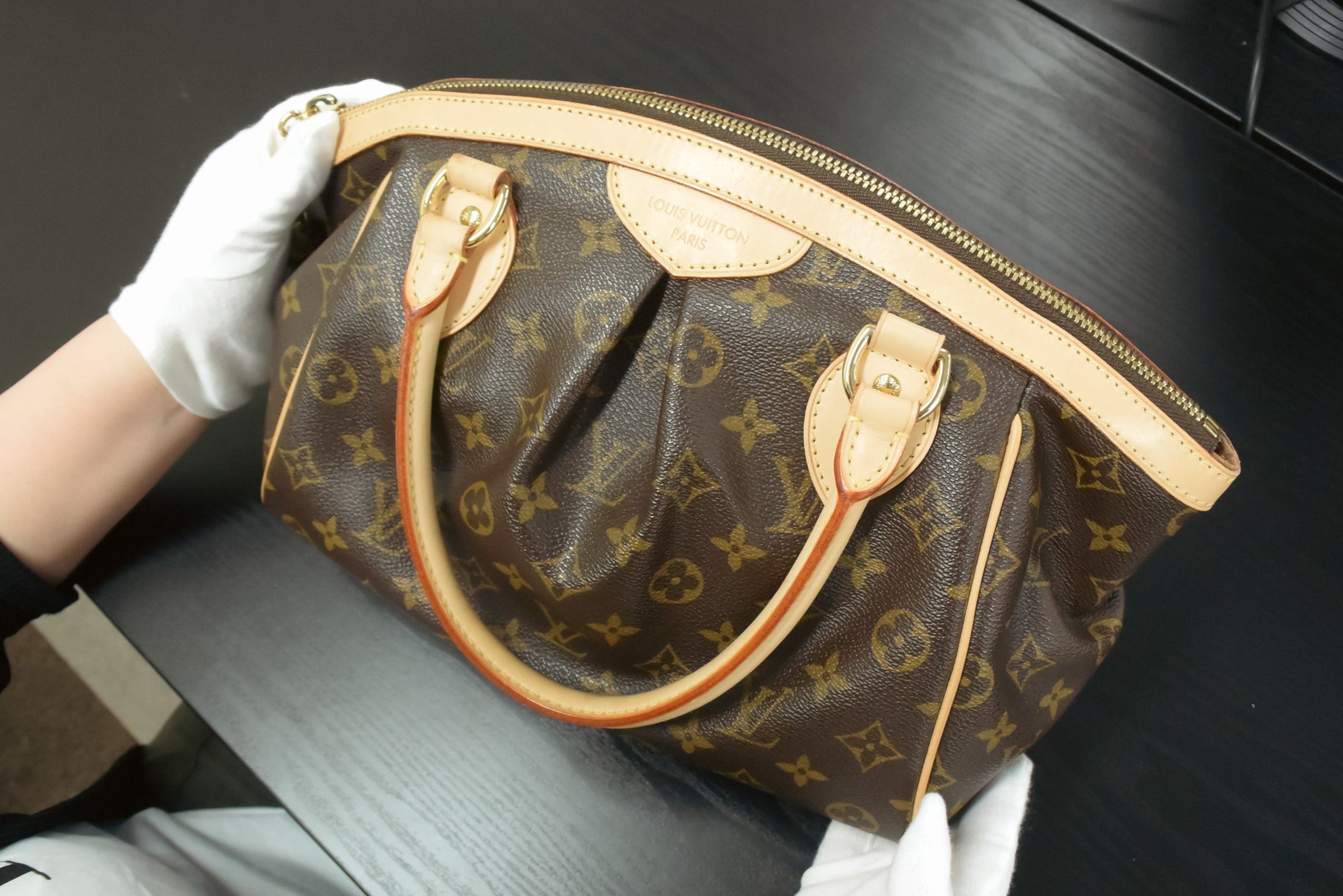 Brown handbag with gold accents held by a gloved hand on a dark surface